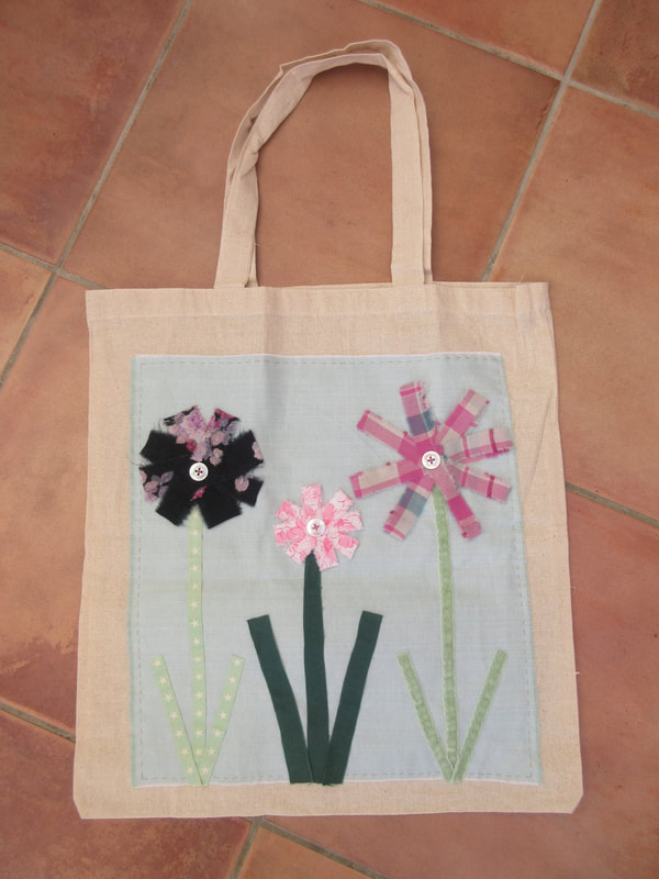 A cream-coloured cloth bag decorated with 3 flowers on stems.  1 flower has a black and pink floral pattern.  1 flower has a pink and white floral pattern.  1 flower has a pink tartan pattern.  All 3 stems are made from green ribbons or strips of green fabric.  There is a button sewn into the centre of each flower.