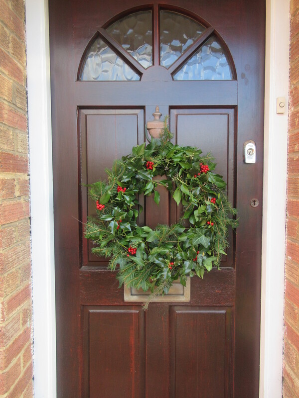 Natural wreath hanging on the front door of a house.  The wreath is mostly sprigs of green holly, ivy and pine but is peppered with red berries from the holly.