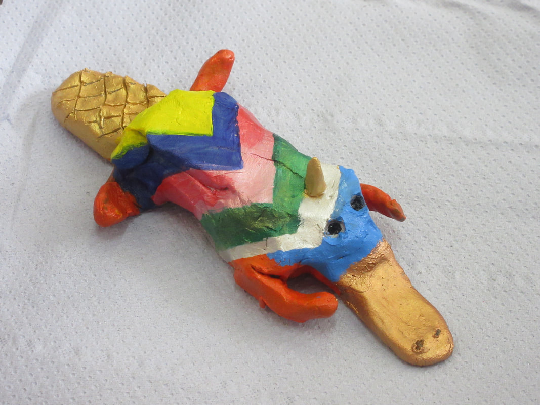 Crude clay model of a duck-billed platypus with a horn on its forehead, like a unicorn.  The body is rainbow striped.  The tail, bill and horn are gold and bronze.