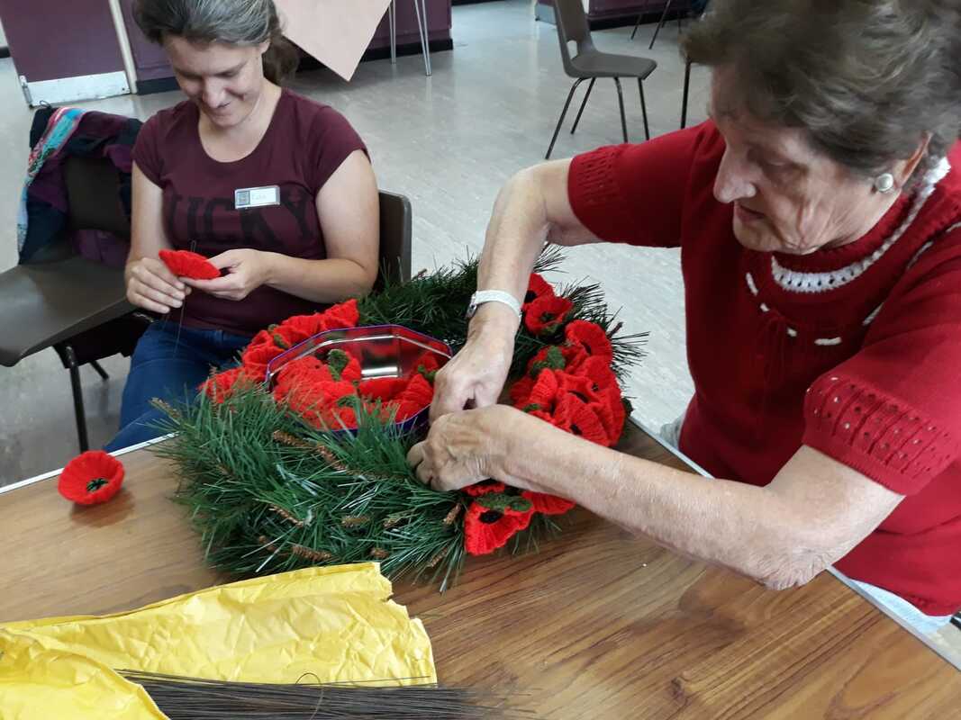 Two women, sitting at a table in a church hall, attaching knitted poppies to a wreath.  One of the women is in her thirties and is wearing a t-shirt with a partially obscured slogan.  The other woman is nearly ninety.