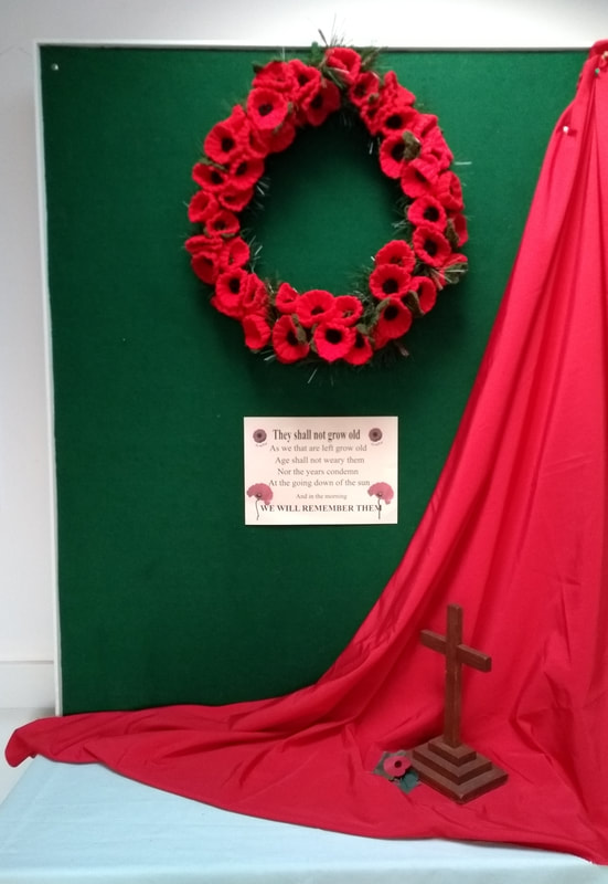 A dark green board decorated with a Remembrance display.  Features include the wreath of knitted poppies, a red cloth draped over one corner, a wooden cross, a traditional paper poppy and a copy of the “They shall not grow old” verse of “For the Fallen” by Laurence Binyon.