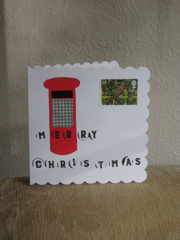 Photo of a white card with a red postbox on it.  The words "Merry Christmas" are written across it so that the E and the R are over the middle of the postbox, like the royal cypher.  There is also a postage stamp with a robin on it.  The card is resting on the arm of a chair.