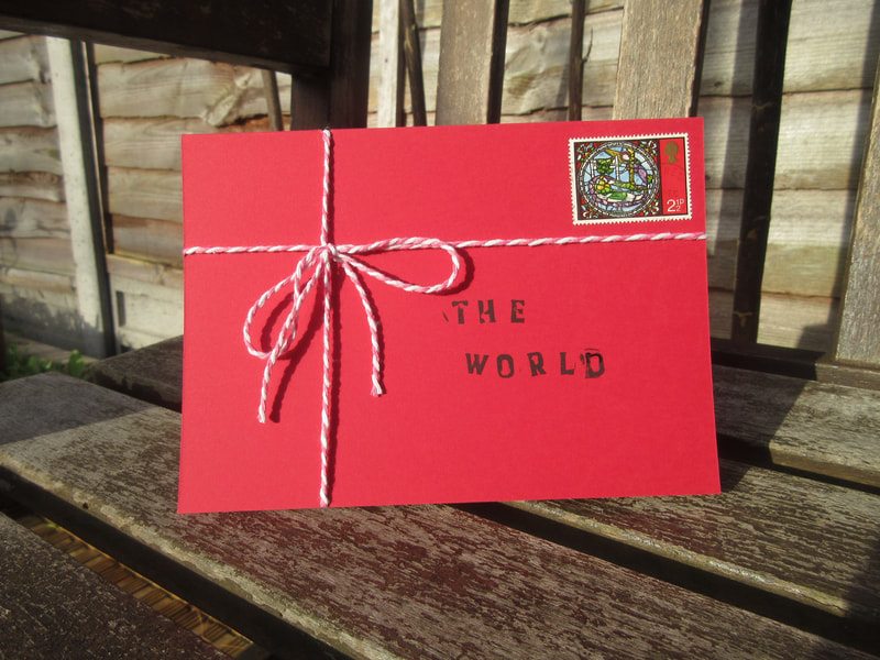 Photo of a red card tied in string, like a parcel.  There is a postage stamp in the top right corner and it is addressed to "The World".  The card is standing on a garden bench in the sunshine.