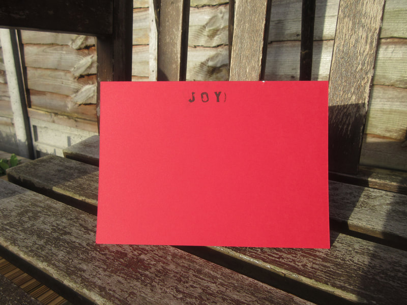 Photo of a red card with "Joy" written in the centre of the top.  The card is standing on a garden bench in the sunshine.