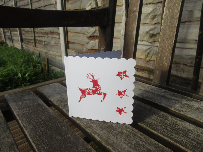 Photo of a white card with scalloped edges.  There is a reindeer and 3 stars on it, all made from red paper with a snowflake pattern.  The card is standing on a garden bench in the sunshine.