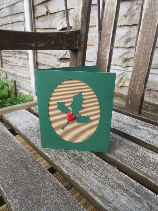 Photo of a green card with an oval cut out to reveal a corrugated cardboard backing.  On the corrogated card is a sprig of green holly leaves and three red berries.  The card is standing on a garden bench in the sunshine.