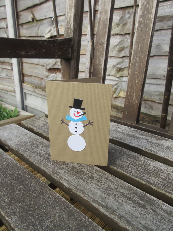 Photo of a buff-coloured card with a snowman on it, made from individual pieces of card.  He wears a black top hat and a blue scarf.  He has a pointy red nose, a big red smile, a black pipe, small black eyes and three black dots for buttons.  His arms look like twigs.  The card is standing on a garden bench in the sunshine.