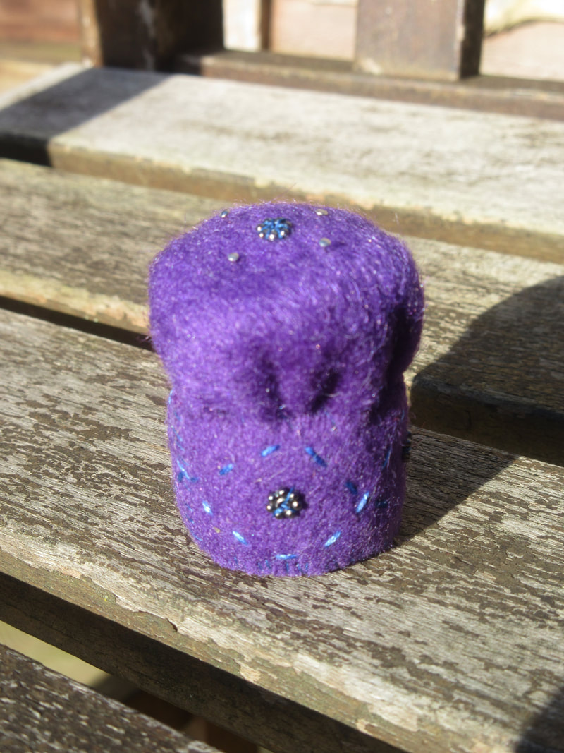 Loosely cylindrical pincushion made of purple felt, embroidered with blue thread and decorated with dark grey metallic beads.