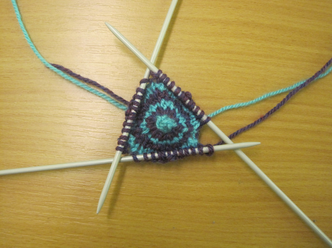 3 double-pointed knitting needles overlapping in a triangle formation.  There are 12 stitches on each needle and a triangle of turquoise and purple striped knitting is suspended between them.