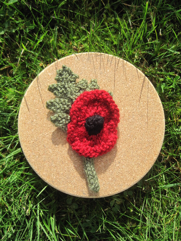 Knitted remembrance poppy with red petals, a black centre and a green leaf and stem.