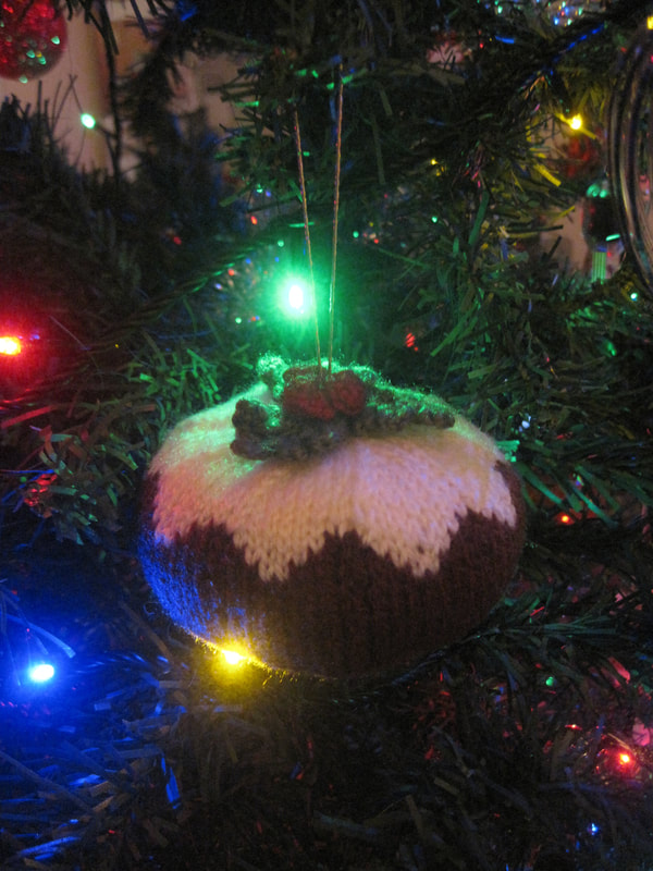 Knitted Christmas pudding hanging from a Christmas tree.  The base of the pudding is brown.  The top is cream with green leaves and red berries attached to it.  The hanging loop is gold.