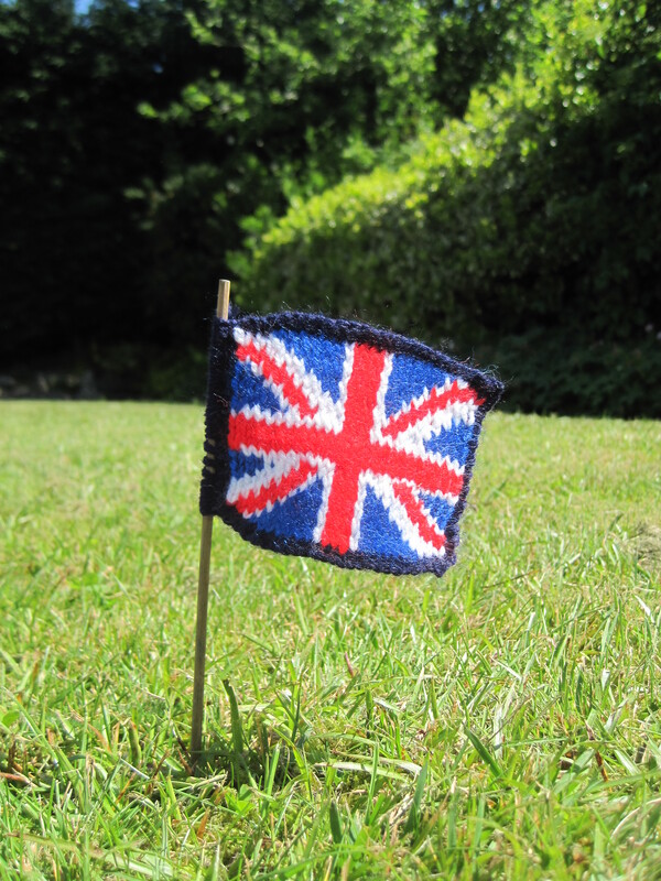 A tiny knitted Union Flag on a tiny pole, sticking out of a garden lawn.
